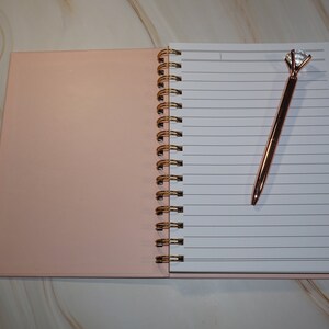 May include: Open notebook with lined pages and a rose gold pen with a diamond-shaped top. The notebook has a pink cover and a gold spiral binding. The pen is resting on the open pages.