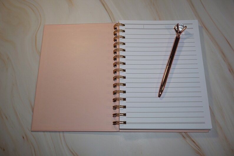 May include: Open notebook with lined pages and a rose gold pen with a diamond-shaped top. The notebook has a pink cover and a gold spiral binding. The pen is resting on the open pages.