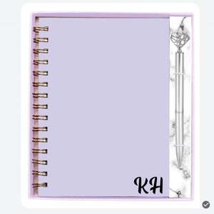 May include: A lavender spiral-bound notebook with gold-colored coils and a matching pen. The pen has a diamond-shaped top and a marble-patterned accent. The initials "KH" are printed on the notebook.