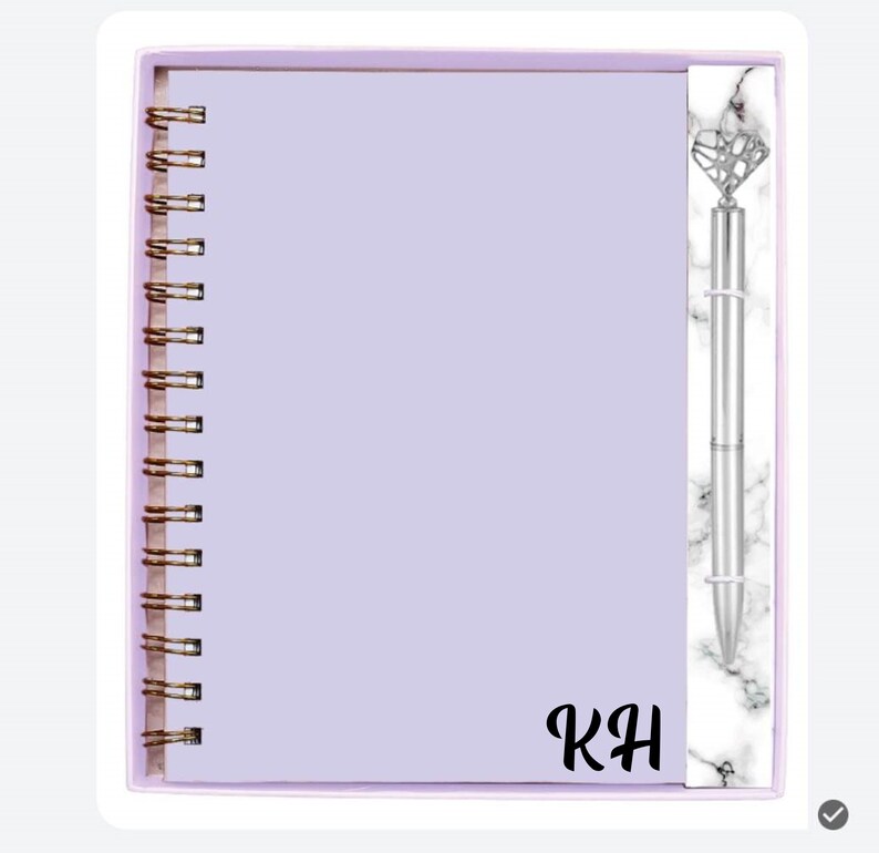May include: A lavender spiral-bound notebook with gold-colored coils and a matching pen. The pen has a diamond-shaped top and a marble-patterned accent. The initials "KH" are printed on the notebook.