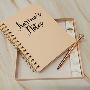 May include: A blush-colored notebook with gold spiral binding and the text "Karina's Notes" in black script. A rose gold pen with a diamond-shaped top rests in the box with the notebook. The set is presented in a matching blush-colored box.