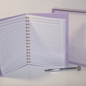 May include: A lavender spiral-bound notebook with lined pages, open to display the interior. A matching box sits behind it, and a silver pen with a crystal-like top rests on the surface. The notebook's binding is gold.