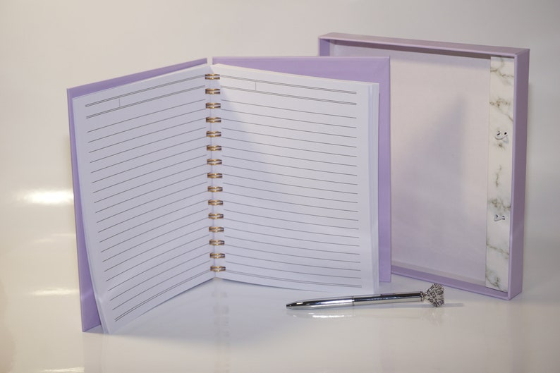 May include: A lavender spiral-bound notebook with lined pages, open to display the interior. A matching box sits behind it, and a silver pen with a crystal-like top rests on the surface. The notebook's binding is gold.