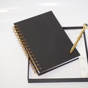 May include: A black spiral-bound notebook with gold-colored coils rests in a black box. A gold pen with a diamond-shaped top is placed inside the box. The notebook and pen are on a white surface.