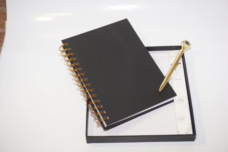 May include: A black spiral-bound notebook with gold-colored coils rests in a black box. A gold pen with a diamond-shaped top is placed inside the box. The notebook and pen are on a white surface.