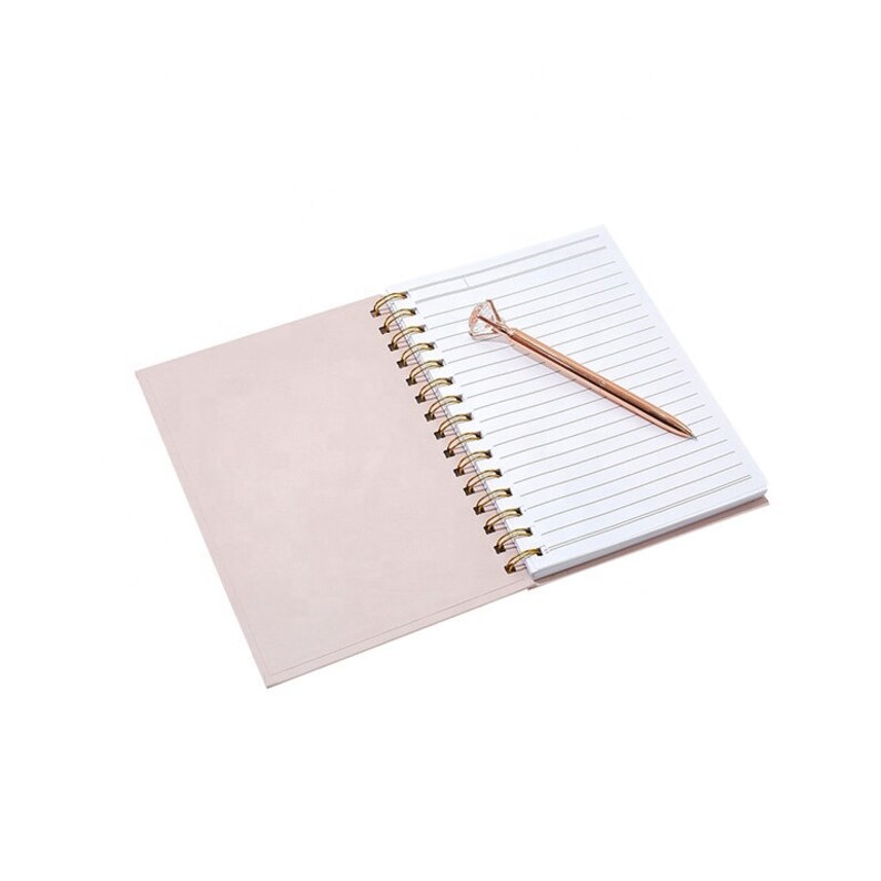 May include: An open spiral notebook with lined pages and a light pink cover. A rose gold pen with a diamond-shaped top rests on the notebook's pages. The notebook's spiral binding is gold.