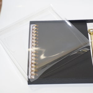 May include: A black spiral-bound notebook and a gold pen with a diamond-shaped top are presented in a black gift box. The pen and notebook are displayed on a white marble-patterned surface. The box is partially covered with a clear plastic sheet.