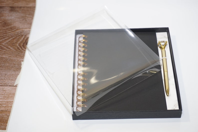 May include: A black spiral-bound notebook and a gold pen with a diamond-shaped top are presented in a black gift box. The pen and notebook are displayed on a white marble-patterned surface. The box is partially covered with a clear plastic sheet.