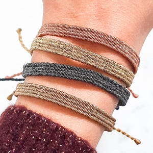 Può includere: Una mano che indossa diversi braccialetti intrecciati nelle tonalità del marrone, oro e grigio. I braccialetti sono impilati sul polso, mettendo in risalto il loro design strutturato. La persona indossa un maglione bordeaux con dettagli dorati.