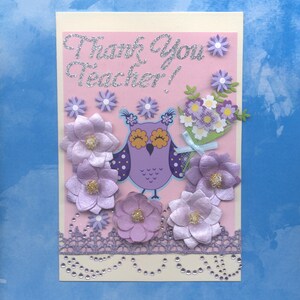 May include: A handmade greeting card with a pink background and the text "Thank You Teacher!" in silver glitter. The card features a purple owl, paper flowers, and a bouquet of flowers. The bottom edge has a decorative lace trim.