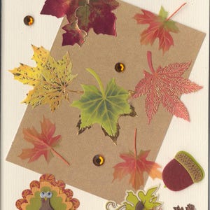 May include: A cream-colored greeting card featuring a brown rectangular paper overlay decorated with various autumn-themed die-cut maple leaves in red, yellow, and green, a felt acorn, a paper turkey, a pumpkin, and amber rhinestones with the text Happy Thanksgiving.