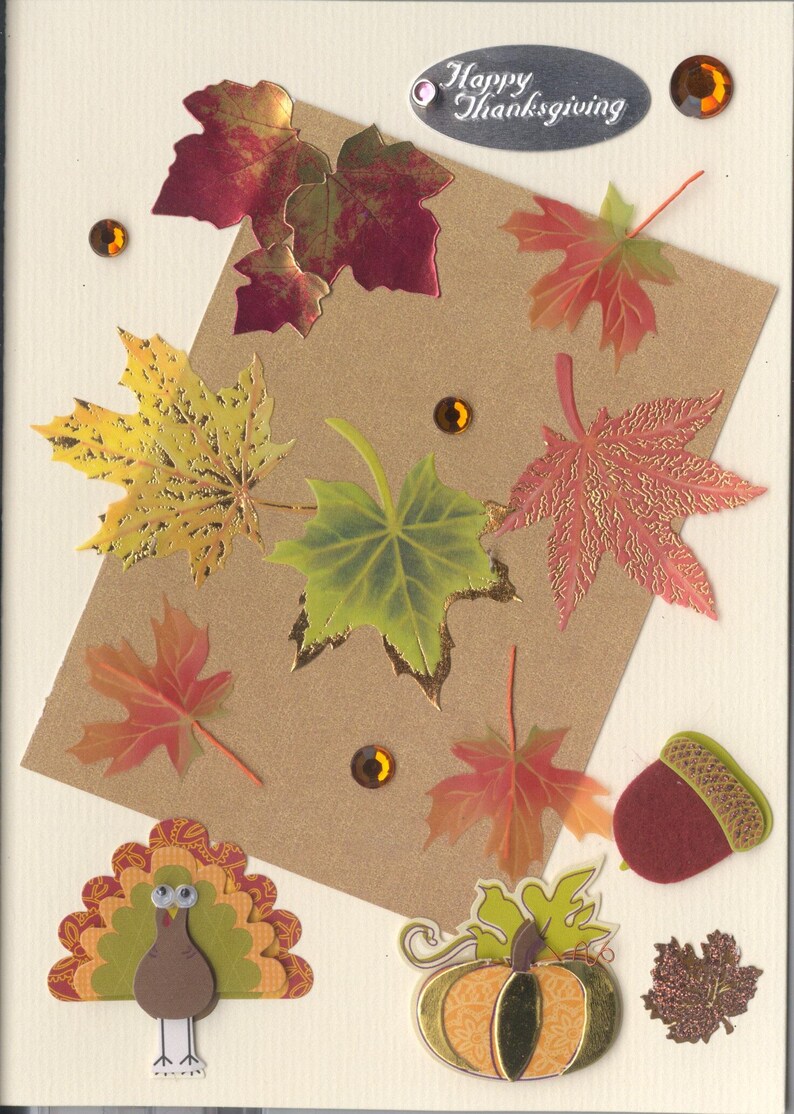 May include: A cream-colored greeting card featuring a brown rectangular paper overlay decorated with various autumn-themed die-cut maple leaves in red, yellow, and green, a felt acorn, a paper turkey, a pumpkin, and amber rhinestones with the text Happy Thanksgiving.