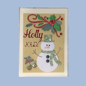 May include: A handmade Christmas card with a glittery gold background. The card features a snowman with a green patterned hat and scarf, and the words "Holly Jolly" in red. Decorative holly and mittens complete the festive design.