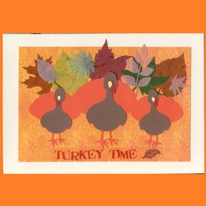 May include: A rectangular greeting card featuring three brown turkey cutouts with red wattles and orange feet. Behind the turkeys are orange cloud shapes and various autumn leaves in shades of green, brown, and purple. The text TURKEY TIME is printed.