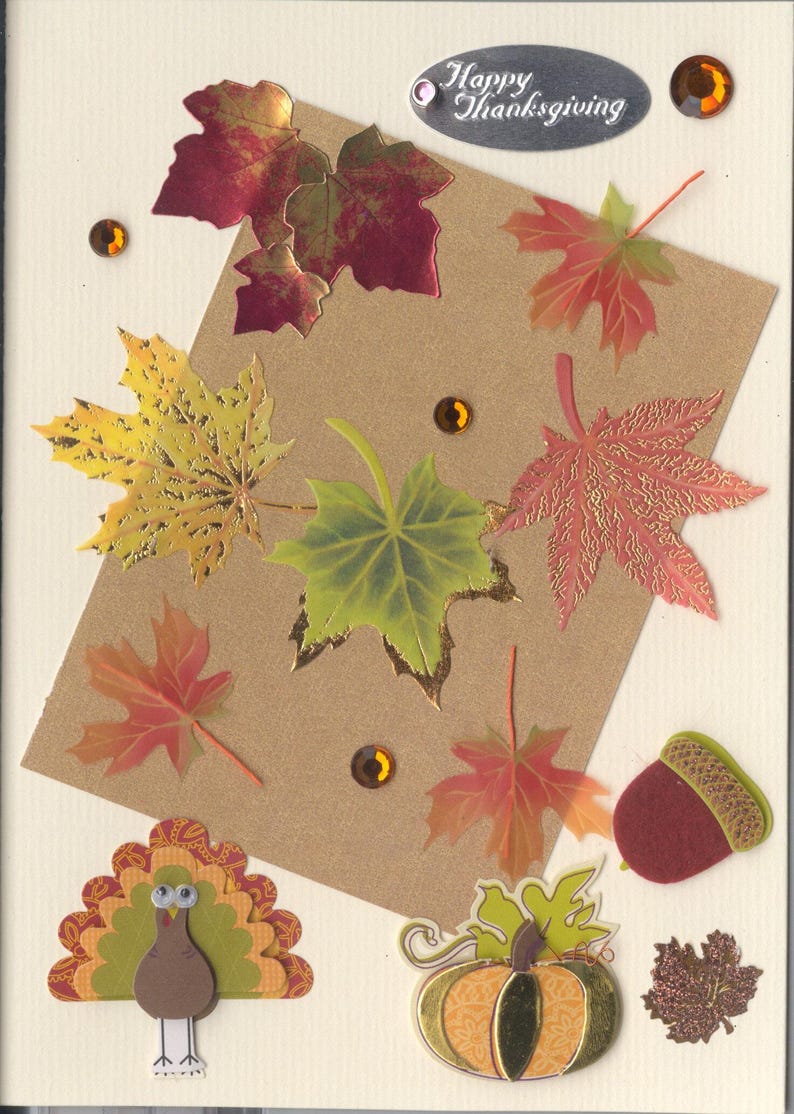 May include: A cream-colored greeting card featuring a brown rectangular paper overlay decorated with various autumn-themed die-cut maple leaves in red, yellow, and green, a felt acorn, a paper turkey, a pumpkin, and amber rhinestones with the text Happy Thanksgiving.