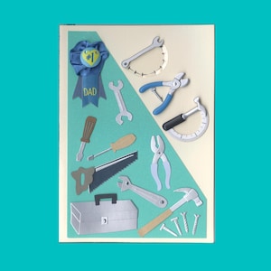 May include: A card with a blue ribbon that says "#1 Dad" and a variety of tools cut out of paper, including a hammer, saw, wrench, pliers, nails, and a toolbox.