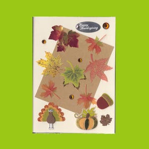 May include: A cream-colored greeting card featuring a brown paper rectangle decorated with various autumn leaf cutouts in red, yellow, and green. The card includes a turkey cutout, a pumpkin, an acorn, small amber rhinestones, and an oval label reading Happy Thanksgiving.