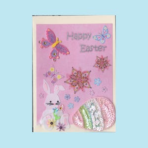 May include: Pink Easter greeting card with glittery text reading "Happy Easter." The card features a felt bunny, butterflies, flowers, and a sequined egg. Decorative elements include glitter and sequins.