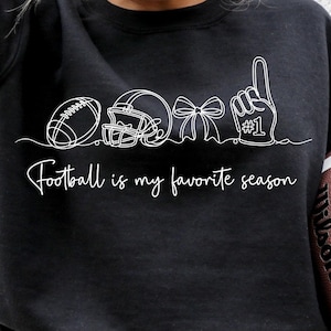 May include: Black sweatshirt with a white line drawing of football-related icons and the text "Football is my favorite season." A brown football is held in the person's hands. The design includes a football, helmet, bow, and foam finger.