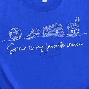 Può includere: Maglietta blu reale con grafica bianca di icone legate al calcio, tra cui un pallone, una scarpa, una porta e un dito di schiuma. Il testo "Soccer is my favorite season" è scritto in corsivo. La maglietta ha anche il testo "DON'T COPY" e "ORIGINAL DESIGN."