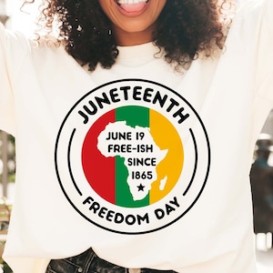 May include: A white t-shirt with a black circle graphic that says "Juneteenth Freedom Day" with a red, green, and yellow outline. The graphic also includes a black silhouette of the continent of Africa with the text "June 19 Free-ish Since 1865" inside the silhouette.