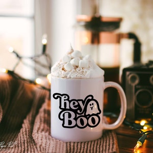 May include: White ceramic mug with a black graphic that says "Hey Boo" with a ghost illustration. The mug is filled with a hot beverage topped with whipped cream and marshmallows.