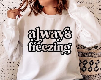 以下が含まれることがあります： 白いスウェットシャツに、レトロなスタイルの黒い文字で「always freezing」と書かれています。モデルはスウェットシャツを着て、模様のあるクッションに座っています。