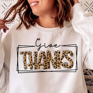 Könnte beinhalten: Weißer Pullover mit einem Leopardenmuster-Druck, auf dem in schwarzer Schrift "Give Thanks" steht.