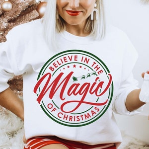 Könnte beinhalten: Weißer Pullover mit einem grünen und roten Kreisdesign, auf dem "Believe in the Magic of Christmas" steht. Das Design enthält auch ein kleines Bild eines von Rentieren gezogenen Schlittens.