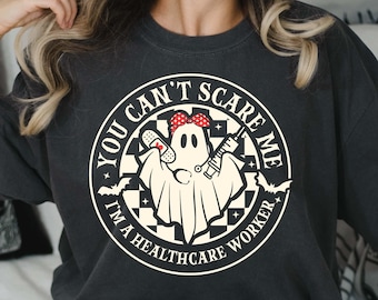 May include: A black t-shirt with a white circle graphic featuring a ghost with a red and white polka dot headband, a bandage, a syringe, and the text "You Can't Scare Me I'm A Healthcare Worker".