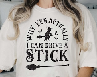 May include: Cream-colored t-shirt with the text "WHY YES ACTUALLY I CAN DRIVE A STICK" in black. The design includes a witch on a broom, bats, and a cat. A broom graphic is below the text.