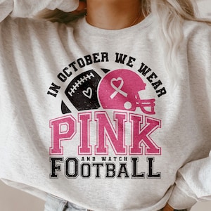 May include: Gray sweatshirt with the text "In October We Wear Pink and Watch Football" in black and pink lettering. A pink ribbon and football helmet are included in the design.