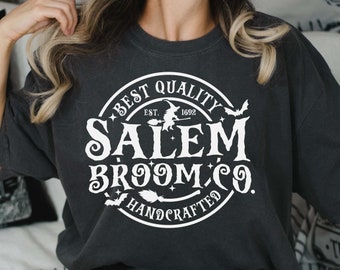 Può includere: Una maglietta grigia scura con una grafica circolare bianca che dice "Best Quality Salem Broom Co. Est. 1692 Handcrafted" con una strega che vola su una scopa e pipistrelli.