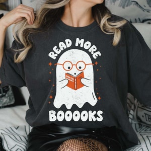 May include: A dark gray t-shirt with a white ghost wearing glasses and reading a book that says "How to Scare" in orange. The text on the shirt says "Read More Booooks" in white.
