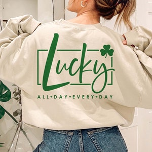 May include: A cream colored sweatshirt with the text "Lucky" in green lettering inside a green rectangle with a green shamrock to the right of the text. The text "ALL DAY EVERY DAY" is written in green below the rectangle.