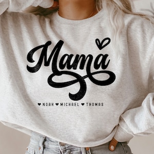 Op de afbeelding: Een grijs sweatshirt met het woord "Mama" in zwarte schrijfletters met een hart erboven. Onder het woord "Mama" staat de tekst "Noah Michael Thomas" met een hart tussen elke naam.
