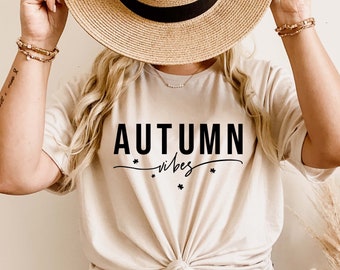 Może przedstawiać: Beżowa koszulka z czarnym napisem "AUTUMN vibes". Koszulka jest zawiązana w pasie. Słomkowy kapelusz trzymany jest nad koszulką. Obraz nawiązuje do tematu jesieni.