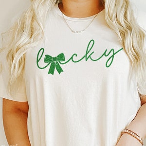 Puede incluir: Una camiseta blanca con la palabra "lucky" escrita en letras de purpurina verde con un lazo de purpurina verde delante de la palabra.