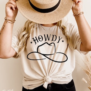 Puede incluir: Una mujer con una camiseta de color crema con un gráfico negro de un sombrero de vaquero y la palabra "HOWDY".