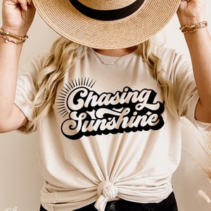Może przedstawiać: Biała koszulka z czarnym nadrukiem w stylu retro, na którym widnieje napis "Chasing Sunshine". Koszulka jest zawiązana w talii, a modelka nosi słomkowy kapelusz.