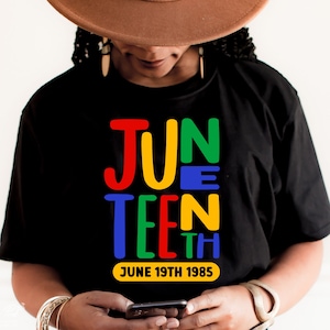 May include: Black t-shirt featuring the word "JUNETEENTH" in red, yellow, green, and blue. The date "JUNE 19TH 1985" is printed below. The person in the image is wearing a brown hat and gold bracelets.