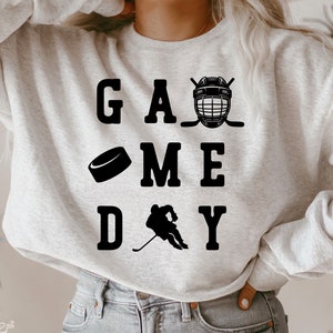 K&ouml;nnte beinhalten: Ein graues Sweatshirt mit dem Text "GAME DAY" in schwarzen Buchstaben. Die Grafik enth&auml;lt einen Hockeyhelm, Hockeyschl&auml;ger, einen Puck und einen Hockeyspieler.