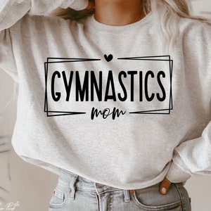 May include: A light gray sweatshirt with the words "GYMNASTICS mom" in black script inside a black rectangular frame. A small black heart is above the word "GYMNASTICS". The sweatshirt is made of a soft material.