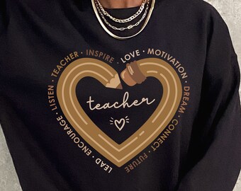 May include: Black sweatshirt with a brown graphic of a pencil shaped into a heart. The heart has the word "teacher" inside and is surrounded by the words "lead, encourage, listen, teacher, inspire, love, motivation, dream, connect, future".