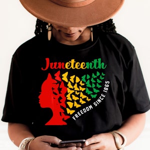 May include: Black t-shirt featuring a red, yellow and green silhouette of a woman's head with butterflies forming a heart shape. The text "Juneteenth Freedom Since 1865" is printed on the shirt.
