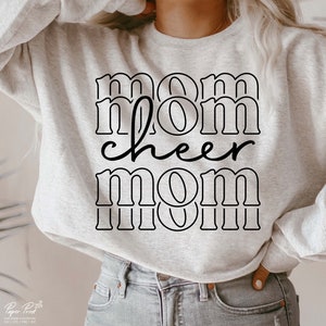 May include: A pale grey sweatshirt featuring the text "mom cheer mom" in black. The word "cheer" is in a cursive font. The sweatshirt is a relaxed fit, ideal for casual wear.