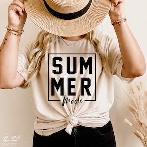 May include: Beige t-shirt with the words "SUMMER MODE" in a black square. The shirt is knotted at the waist. A straw hat is held above the shirt. The image also shows a person wearing bracelets.