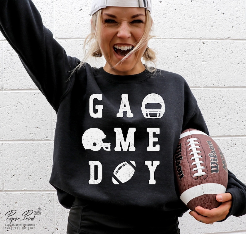May include: A black sweatshirt with white text that reads "GAME DAY" with football and helmet graphics. The person wearing the sweatshirt is holding a brown football.