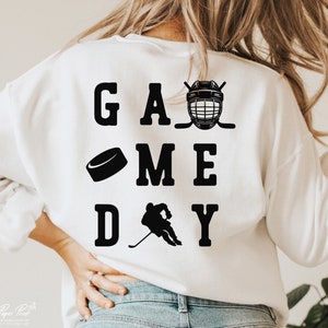 K&ouml;nnte beinhalten: Wei&szlig;er Pullover mit schwarzem Text, der "GAME DAY" lautet, mit einem Hockeyschl&auml;ger, einem Puck und einer Hockeyfigur.