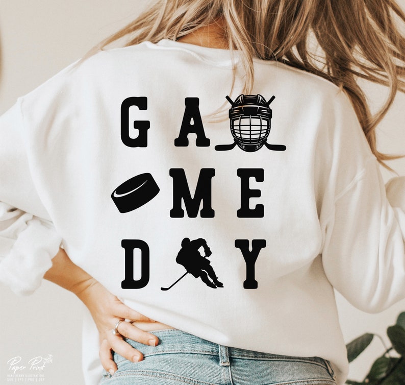 K&ouml;nnte beinhalten: Wei&szlig;er Pullover mit schwarzem Text, der "GAME DAY" lautet, mit einem Hockeyschl&auml;ger, einem Puck und einer Hockeyfigur.